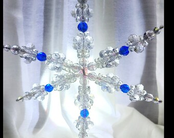 Pode incluir: Um enfeite de floco de neve de cristal com contas azuis. O floco de neve é feito de contas de vidro transparentes e tem um laço de arame prateado para pendurar.