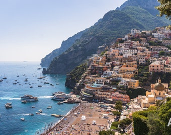 Può includere: Una vista panoramica di Positano, in Italia, una città colorata costruita su una scogliera scoscesa che si affaccia sul Mar Mediterraneo. La città è piena di edifici in tonalità di giallo, arancione, rosa e bianco, con una chiesa in fondo alla collina. Il mare è di un blu profondo e ci sono molte barche in acqua.