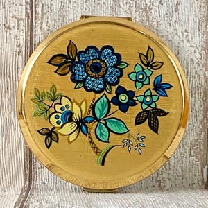 May include: A vintage, round compact with a gold finish. The compact features a floral design in shades of blue, green, and yellow. The flowers and leaves are arranged on a gold background, creating a decorative and elegant look.