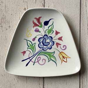 May include: A white, triangular ceramic plate featuring a hand-painted floral design. The flowers are in blue, red, yellow, and purple, with green leaves and stems. The plate has a slightly raised edge and a matte finish.