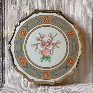 May include: A gold-toned compact mirror with a white porcelain lid featuring a floral design. The design depicts a teapot with pink flowers and green leaves on a green pedestal. The border of the mirror is gold with a blue and brown pattern.