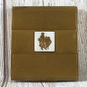 Op de afbeelding: Een gouden compact met een wit emaille ontwerp met een groep mensen in een scène. De compact heeft een getextureerd oppervlak.