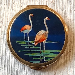 May include: A round, vintage compact with a gold-tone metal frame. The compact features a painted scene of two flamingos in a blue water setting with green lily pads. The flamingos have pink and orange plumage.