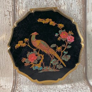 May include: A round, gold-toned metal compact mirror featuring a black surface decorated with a colourful illustration of a long-tailed bird perched on a flowering branch. The design includes pink and red blossoms with green leaves and gold accents.