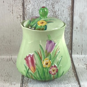 Könnte beinhalten: Ein hellgrüner Keramiktopf mit Deckel, verziert mit farbenfrohen floralen Designs. Der Topf zeigt geprägte Tulpen, Narzissen und andere Blumen mit grünen Blättern und Stielen. Der Deckel hat einen grünen Knopf.