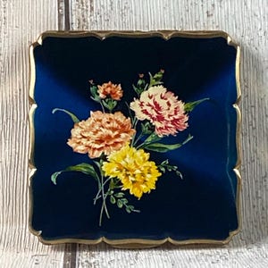May include: A vintage blue and gold compact with a floral design. The flowers are yellow, pink, and orange and are painted on a dark blue background. The compact has a gold rim.