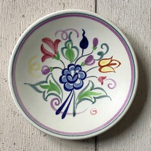 May include: A small, round, white ceramic dish with a hand-painted floral design. The design features a central blue flower surrounded by pink, yellow, green, and purple flowers and leaves. The rim has a lavender and green border.