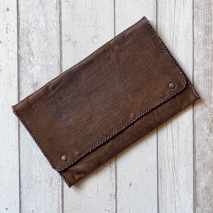 Op de afbeelding: Een bruine leren clutch met een krokodillenstructuur. De clutch heeft een klepsluiting met twee zilverkleurige klinknagels en is aan de randen gestikt met een donkerdere bruine draad. De clutch is rechthoekig en lijkt gesloten.