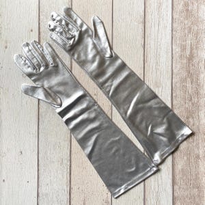 Puede incluir: Un par de guantes largos de ópera que llegan hasta el codo, con un acabado en color plata metálico. Los guantes presentan una superficie lisa y reflectante con un diseño ajustado que se extiende más allá de la muñeca hasta el antebrazo.