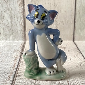 May include: A ceramic figurine of Tom Cat from the Tom and Jerry cartoon series. The figure is light blue with white accents on the chest and belly. The cat is posed leaning against a green base.