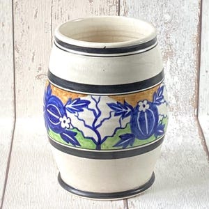 Könnte beinhalten: Eine cremefarbene Keramikvase mit zylindrischer Form. Die Vase hat schwarze Streifen oben und unten und ein Blumendesign in Blau, Grün und Gold in der Mitte. Das Blumendesign umfasst blaue Blumen und Blätter.