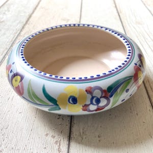 Peut inclure: Un bol rond en céramique avec un intérieur beige clair lisse et un extérieur blanc orné d'un motif floral peint à la main. Le design comprend des fleurs jaunes, rouges et violettes avec des feuilles vertes, soulignées par un bord bleu pointillé.