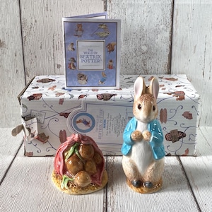 May include: A ceramic Peter Rabbit figurine with a blue jacket, next to a ceramic onion bag and a box with illustrations. The box and the book in the background have the text "THE WORLD OF BEATRIX POTTER".