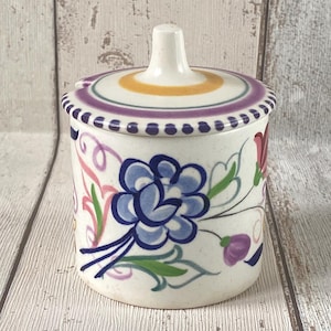 Puede incluir: Un azucarero de cerámica con tapa. El cuenco es blanco con un diseño floral en azul, rosa, morado y verde. La tapa tiene un asa y está decorada con rayas amarillas, moradas y azules. El borde de la tapa tiene un patrón de puntos.