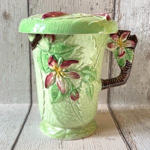 Puede incluir: Una taza de cerámica verde claro con tapa a juego. La taza presenta un diseño floral con flores rojas y blancas y hojas verdes. El asa es marrón y tiene un adorno floral. La taza mide aproximadamente 15 cm de alto.