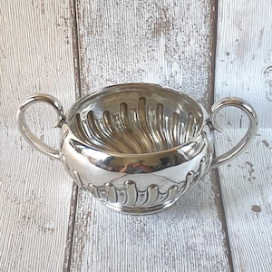 May include: A polished silver-tone sugar bowl featuring two curved handles on opposite sides. The exterior and interior surfaces are decorated with a repeating vertical fluted pattern that extends from the base towards the rim of the bowl.
