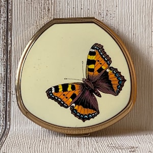 May include: A gold-toned compact with a white background and a colourful butterfly design. The butterfly has orange, black, and white wings.