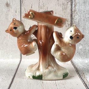 May include: A ceramic figurine featuring two brown and white bears climbing a central brown tree trunk. The bears have painted black eyes and noses, and the tree base includes small patches of green paint to represent grass at the bottom.