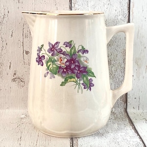 May include: Cream-colored ceramic pitcher with a gold rim and handle. A floral design of purple and white violets with green leaves decorates the front. The pitcher has a classic, rounded shape, suitable for serving beverages.