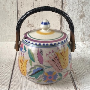 Könnte beinhalten: Eine Zuckerdose aus Keramik mit Deckel und geflochtenem Griff. Die Schale ist mit bunten Blumenmustern in Gelb, Rosa, Blau und Grün verziert. Der Deckel hat einen gestreiften Knauf und einen lila und grünen Rand.