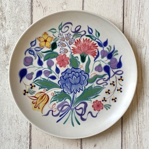 Può includere: Un piatto in ceramica bianca con un motivo floreale dipinto a mano. Il design presenta un bouquet di fiori nei toni del blu, rosa, giallo e viola, con foglie verdi e nastri viola. Il piatto è rotondo e ha un bordo semplice.