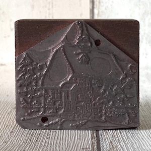 May include: A vintage metal stamp depicting a house and trees, set on a dark brown wooden block. The metal stamp has a detailed illustration of a house with a peaked roof and surrounding trees. The metal surface has a weathered appearance, suggesting age and use.