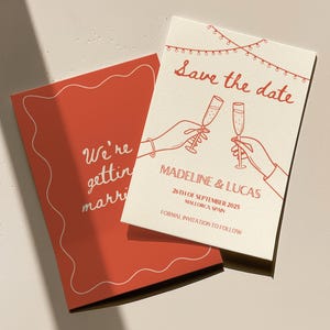 以下が含まれることがあります： 赤と白のテキストで書かれた、結婚式の招待状。シャンパンのグラスで乾杯するカップルと、「Save the date」の文字、Madeline & Lucasの名前、2025年9月26日、スペインのマヨルカ島、そして「正式な招待状は後日」という文字が書かれている。