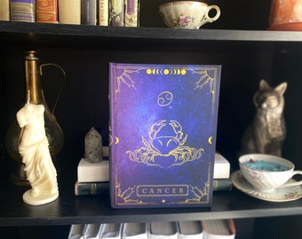 Puede incluir: Un diario de tapa dura azul y dorado con un diseño celestial que presenta un cangrejo y la palabra "CANCER".