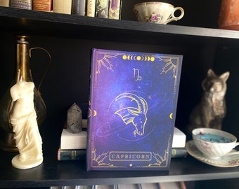 Puede incluir: Un diario azul y dorado con un diseño de galaxia y el símbolo del zodíaco de Capricornio. El texto "CAPRICORN" está impreso en dorado en la portada.