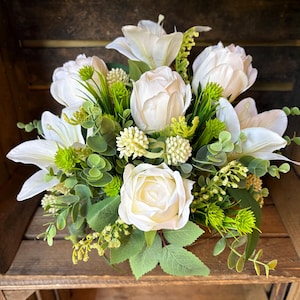 May include: A floral arrangement featuring white lilies, tulips, and roses interspersed with green eucalyptus leaves and small, textured lime green filler flowers. The bouquet is densely packed with various botanical elements in shades of white and green.