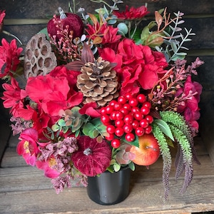 May include: A floral arrangement in a black pot featuring red velvet pumpkins, a large pinecone, bright red berries, a red apple, dried lotus pods, and various red silk flowers with green foliage, creating a dense, multi-textured seasonal display.
