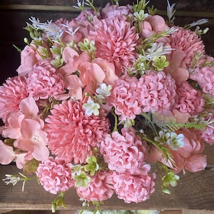 May include: A vibrant bouquet of artificial flowers in shades of pink and peach. The arrangement includes large, spherical blooms and smaller, clustered flowers, with touches of white and green foliage. The flowers are arranged in a dense, full composition.