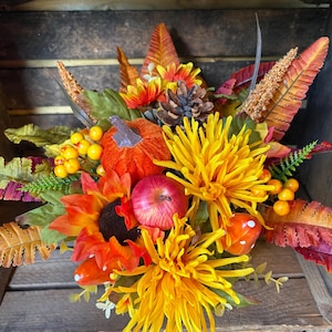 May include: A decorative autumn floral arrangement featuring bright yellow chrysanthemums, an orange fabric pumpkin, a red apple, a brown pinecone, orange sunflowers, green fern leaves, yellow berries, and small red mushrooms with white spots.
