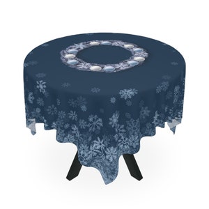以下が含まれることがあります： 白い雪の結晶模様の青い丸いテーブルクロス。テーブルクロスの中央には、白いオーナメントが付いたリースがあります。