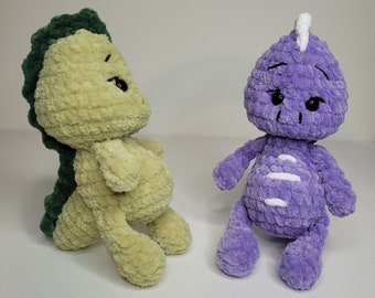May include: Two crocheted stuffed dinosaur toys. One is green and yellow, the other is purple. Both have black eyes and white details.