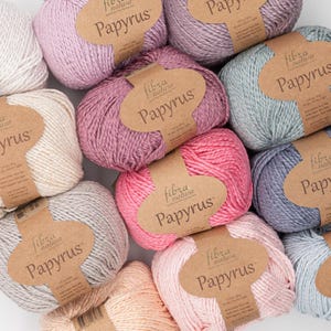 May include: A collection of colorful yarn balls in various shades of pink, purple, gray, and blue. Each ball has a brown paper label with the text "fibra natura Papyrus". The yarn is tightly wound and appears soft.