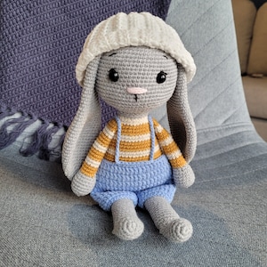 May include: A gray crocheted bunny wearing a white crocheted hat and blue overalls with yellow stripes. The bunny is sitting on a gray fabric surface.