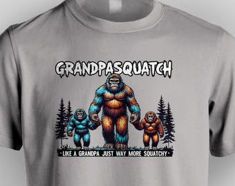 May include: A gray t-shirt with a graphic of three Bigfoot figures. The largest figure is in the center and has orange fur with blue accents. The two smaller figures are on either side of the larger figure and have blue fur. The text "Grandpasquatch" is above the figures and the text "Like a grandpa just way more squatchy" is below the figures.