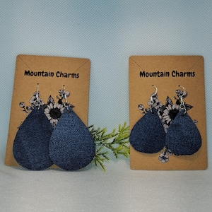 May include: Two pairs of dark blue denim teardrop earrings on display cards. The cards are brown with the text "Mountain Charms" and floral accents. The earrings hang from silver hooks.