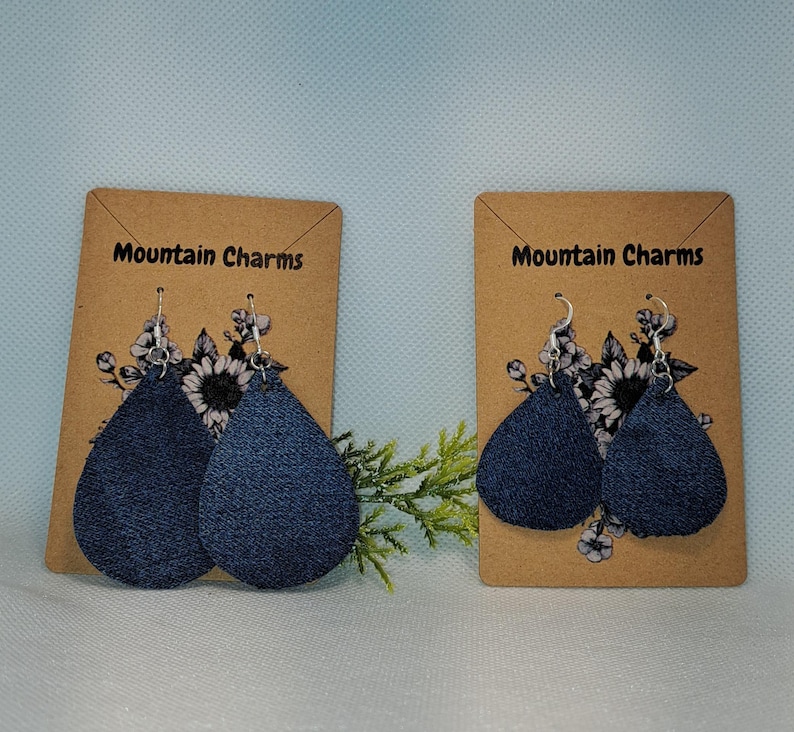 May include: Two pairs of dark blue denim teardrop earrings on display cards. The cards are brown with the text "Mountain Charms" and floral accents. The earrings hang from silver hooks.