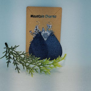 May include: Teardrop-shaped earrings with a denim-like texture, featuring a light-colored floral design at the top. The earrings hang from silver-colored hooks. The product is displayed on a brown card with the text "Mountain Charms".