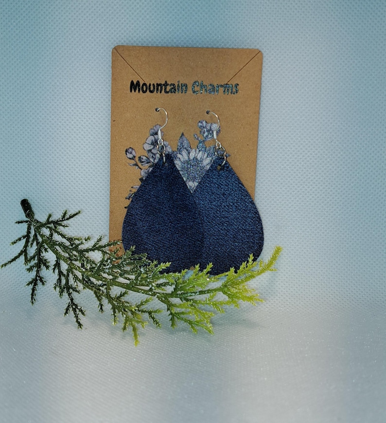 May include: Teardrop-shaped earrings with a denim-like texture, featuring a light-colored floral design at the top. The earrings hang from silver-colored hooks. The product is displayed on a brown card with the text "Mountain Charms".