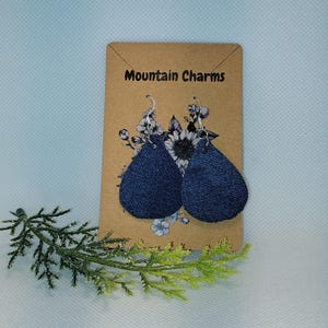 May include: A pair of dark blue teardrop-shaped earrings with silver hooks. The earrings are displayed on a brown card with the text "Mountain Charms" and floral accents. A sprig of greenery is in the foreground.