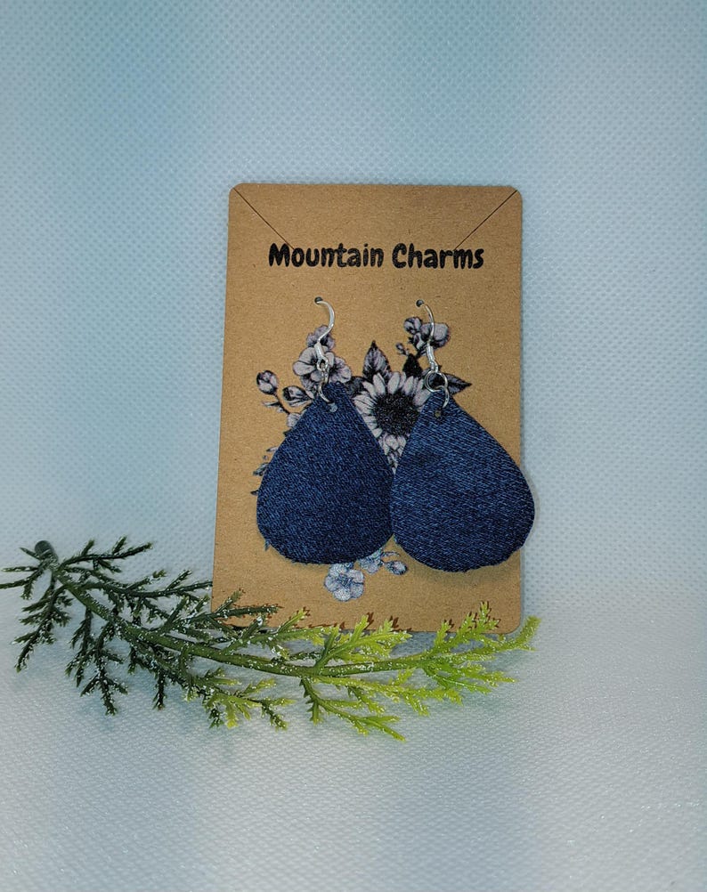 May include: A pair of dark blue teardrop-shaped earrings with silver hooks. The earrings are displayed on a brown card with the text "Mountain Charms" and floral accents. A sprig of greenery is in the foreground.