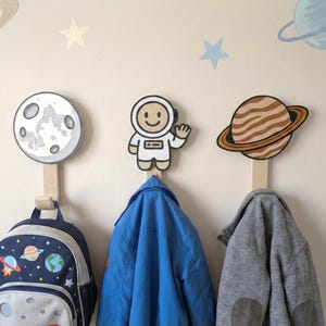 May include: Three space-themed wall hooks with a moon, astronaut, and planet design. A blue backpack and two jackets hang from the hooks. The hooks are mounted on a light beige wall with space-themed wall decals.