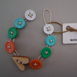 May include: A colorful button bracelet featuring white, turquoise, green, and orange buttons strung together with white thread. A wooden heart-shaped button serves as a clasp. The tag reads "McSickCreations.Etsy.com" and the bracelet is 6 1/2 inches long.