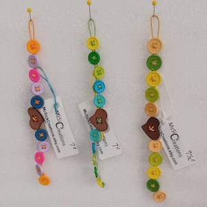 May include: Three colorful button bracelets with wooden heart charms. Each bracelet features a different color scheme, including pink, blue, green, yellow, and orange. The bracelets are strung on a thin cord and have a small loop at the top for hanging.