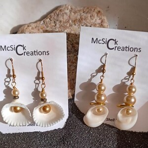 May include: Two pairs of gold tone earrings with white seashells and pearls. The earrings on the left have a single pearl and a seashell. The earrings on the right have three pearls and a seashell. The card reads "McSick Creations".