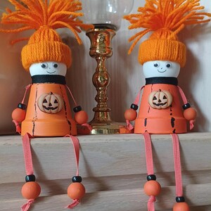 May include: Two decorative figures crafted from orange terracotta pots, each wearing a bright orange yarn hat with a pom-pom top. The figures feature white heads with googly eyes, a black collar, and a small pumpkin emblem on the chest.