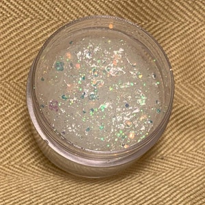 Puede incluir: Un tarro transparente lleno de brillo de labios blanco y brillante. El brillo de labios tiene brillo iridiscente en todo.